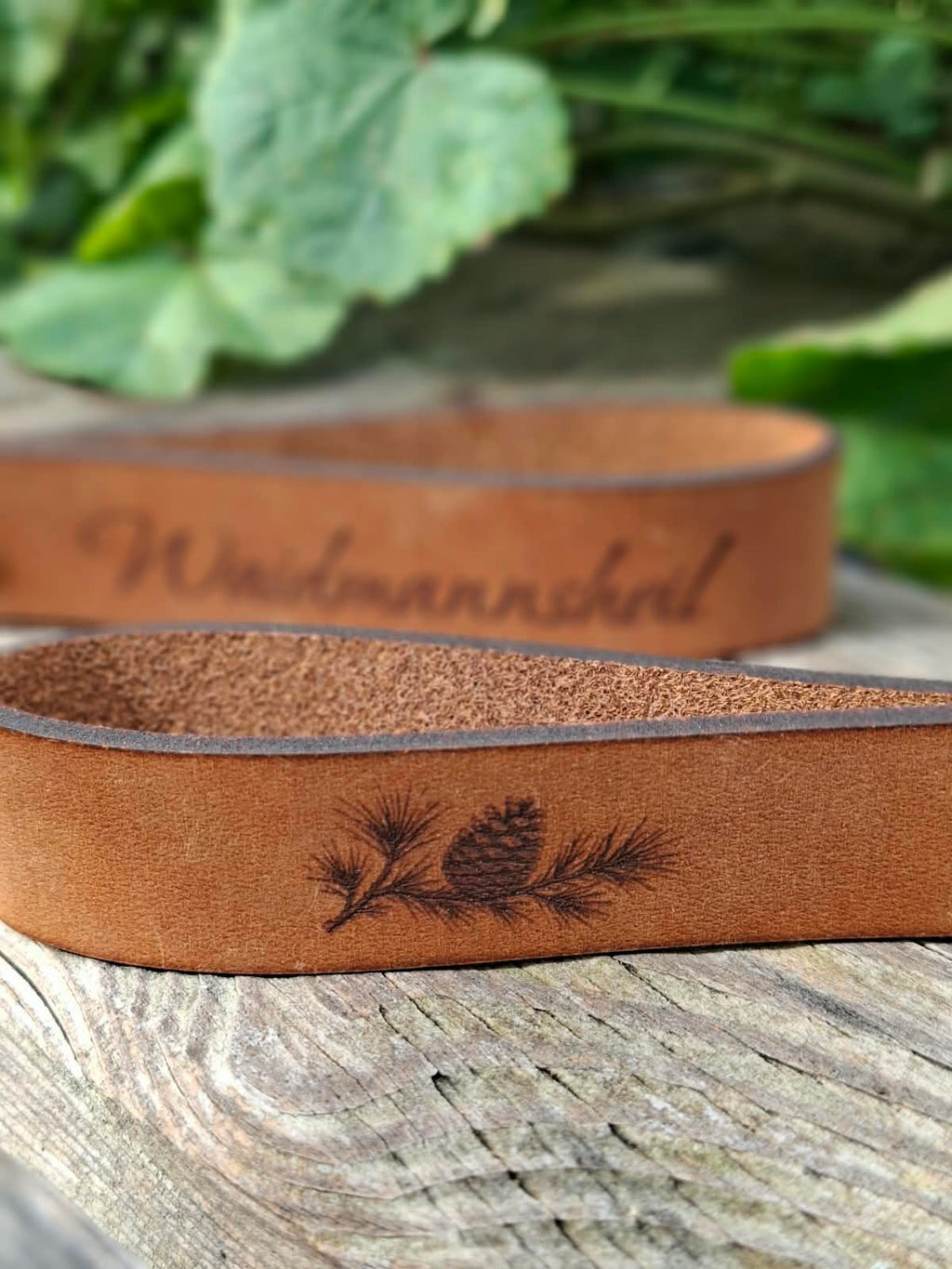 Leder-Schlüsselanhänger mit Jagdmotiv auf einem Holztisch neben grünen Blättern
